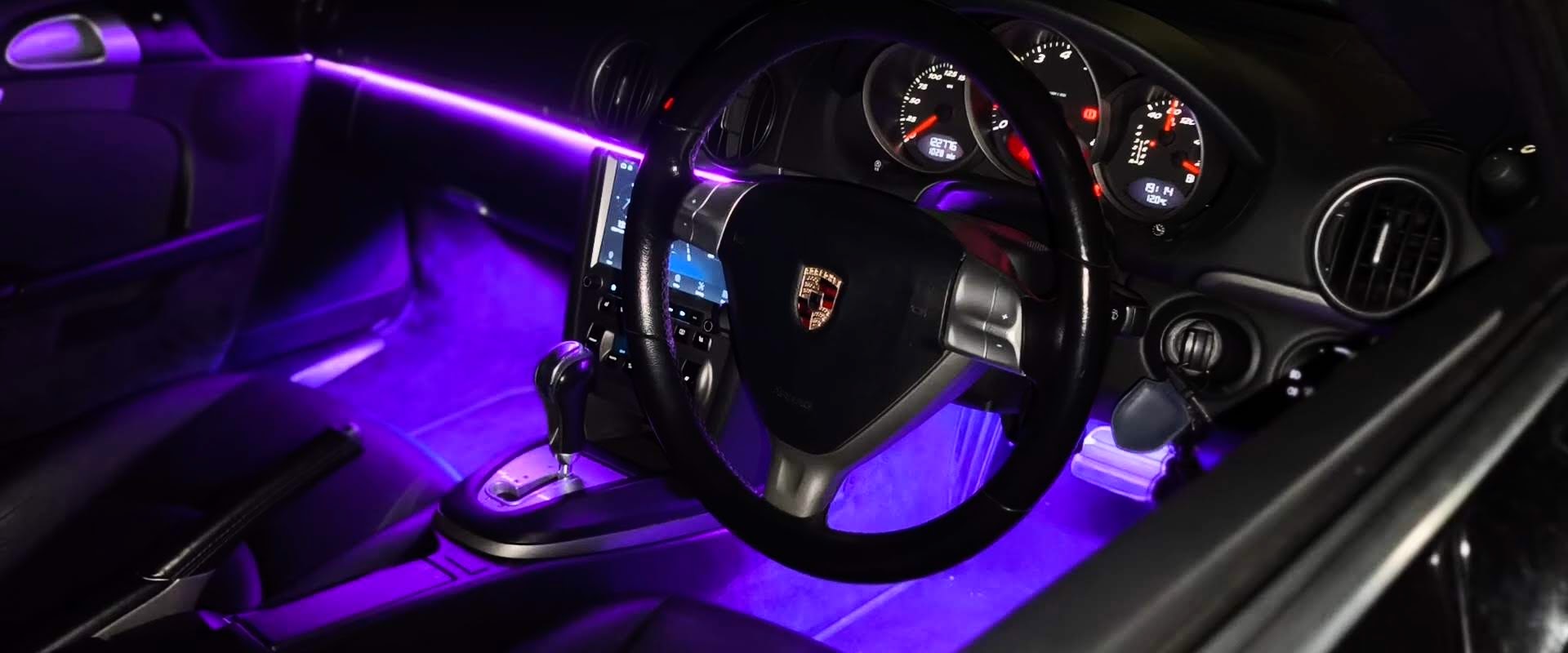Car interior with steering wheel and dashboard illuminated by purple LED lights.