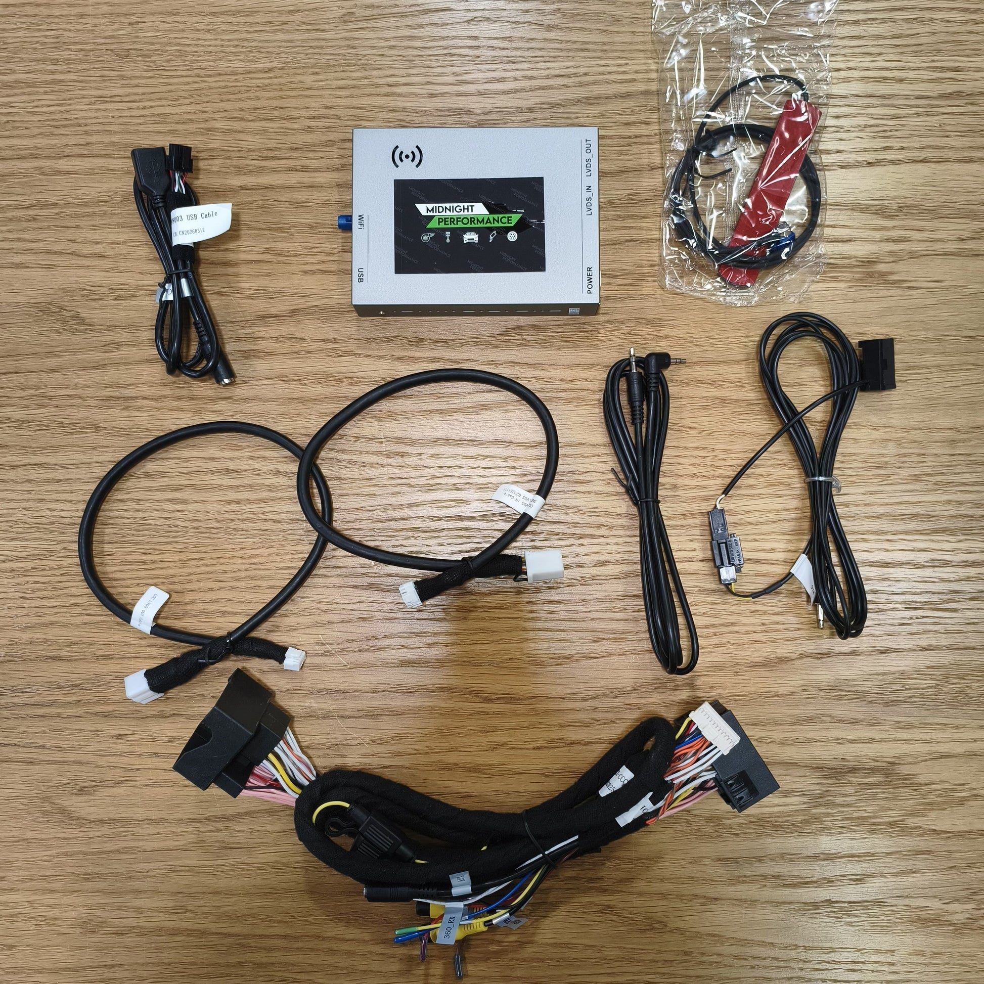 An automotive wiring harness kit laid out on a wooden table, including a central white control module with a screen, multiple black cables with various connectors, a harness bundle with multicolored wire colors, and a small packaged red and black accessory.
