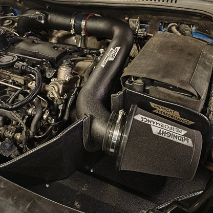 A black aftermarket car air intake system with a conical filter and corrugated tube, installed in a vehicle engine bay. The filter has a black foam cover with a white 'MIDNIGHT PERFORMANCE' logo.