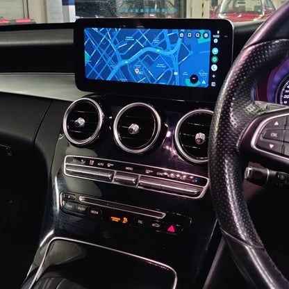 A close-up view of a car's dashboard and center console, showing a large touchscreen infotainment display with a blue map, three circular air vents, and a black steering wheel in a garage setting.