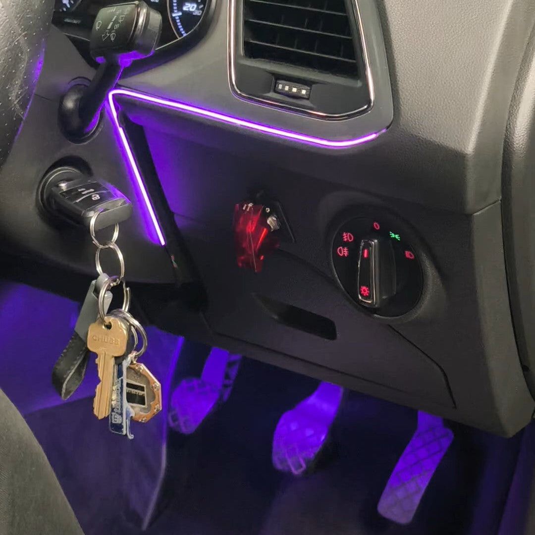 A close-up of a car's dashboard and center console showing a purple ambient light strip under the air vents and around the ignition. A set of keys hangs from the keyhole, and a person's white sneakers are visible at the bottom edge.
