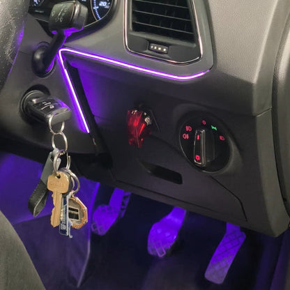 A close-up of a car's dashboard and center console showing a purple ambient light strip under the air vents and around the ignition. A set of keys hangs from the keyhole, and a person's white sneakers are visible at the bottom edge.