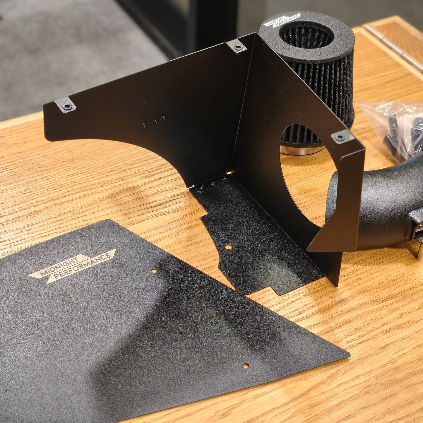 Black metal intake shield kit with a black air filter and black hose, displayed on a wooden table with a branded splash shield in the foreground.